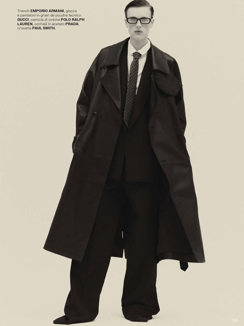 Full-length portrait of an adult male wearing a black suit, trench coat, eyeglasses, and tie in a fashion editorial photo.