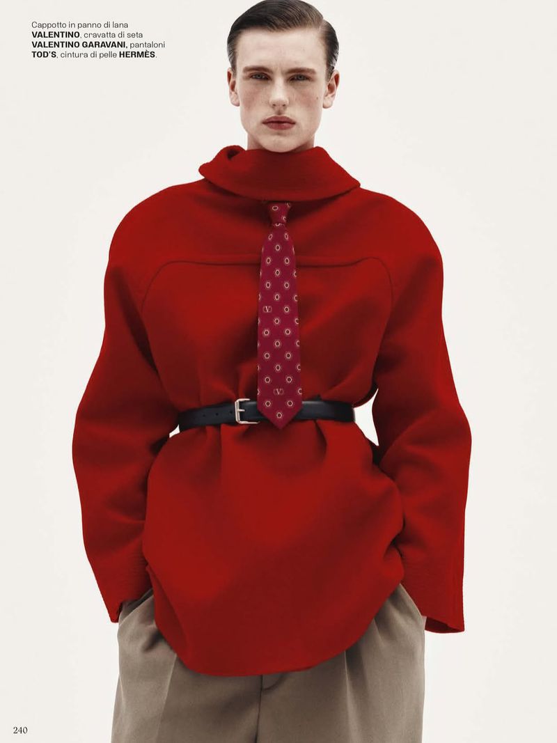 Editorial fashion shot of a male model in an oversized red turtleneck sweater, black belt, and patterned tie against a neutral backdrop.
