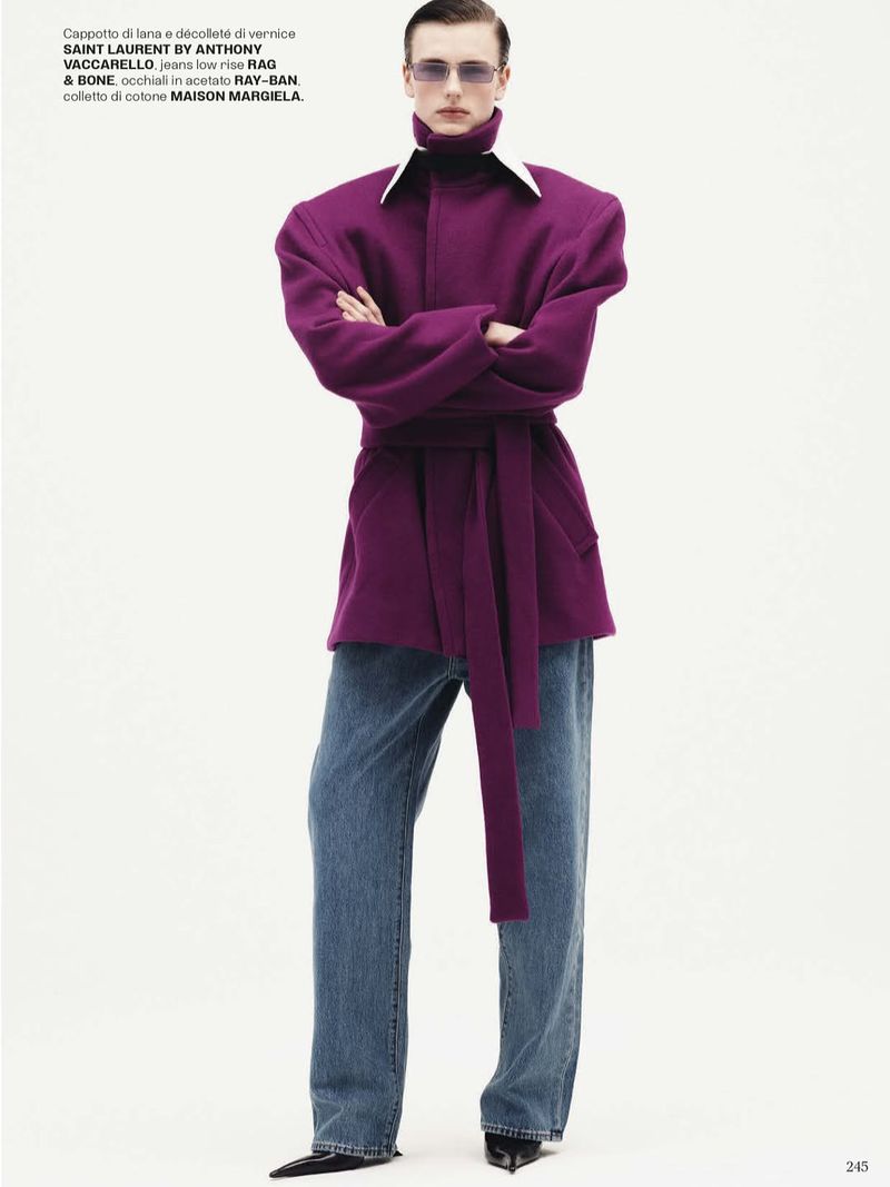 Fashion shot of a man wearing purple coat with jeans and sunglasses, standing against a white background in a studio setting.