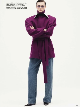 Fashion shot of a man wearing purple coat with jeans and sunglasses, standing against a white background in a studio setting.