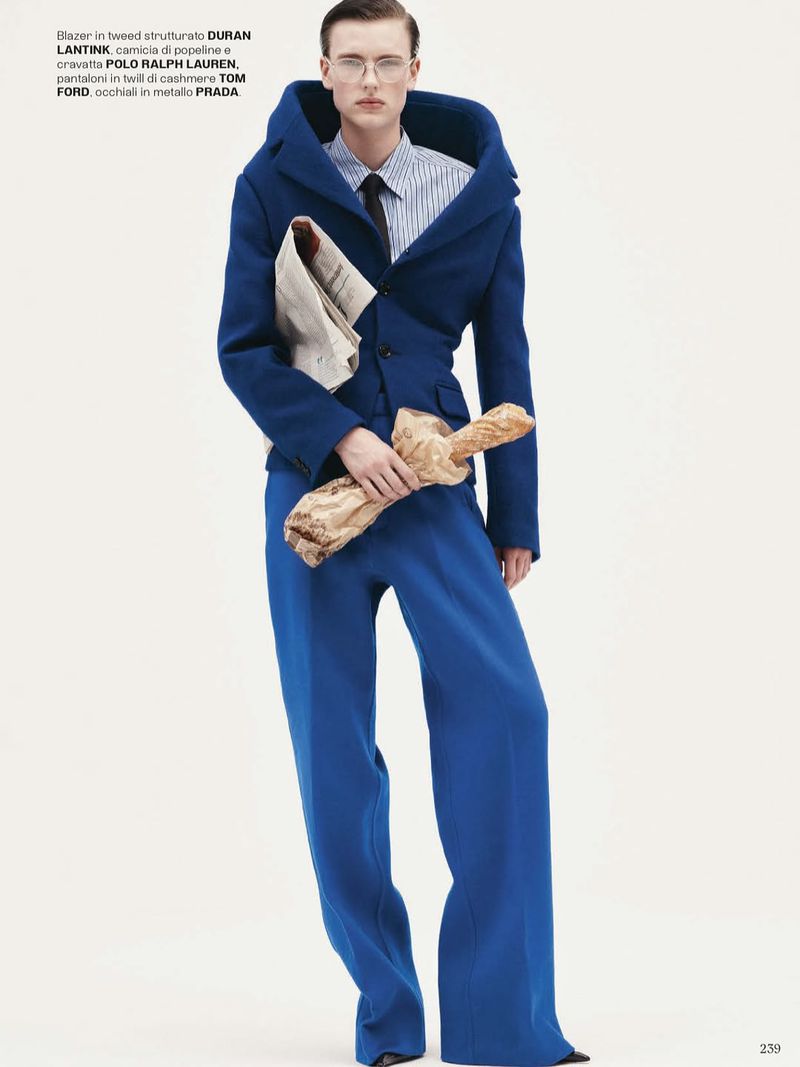 Fashion model wearing a blue blazer and matching wide-leg pants stands in a studio holding a newspaper and loaf of bread.