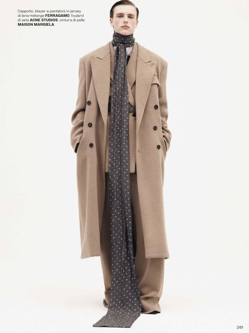 A slender man is standing in a full shot, showcasing a stylish tan overcoat, blazer, trousers, and a long patterned scarf against white.