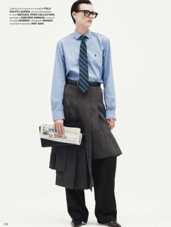 Editorial fashion shot of a young adult male with black hair wearing glasses, a light blue shirt, tie, grey skirt, and carrying newspaper.