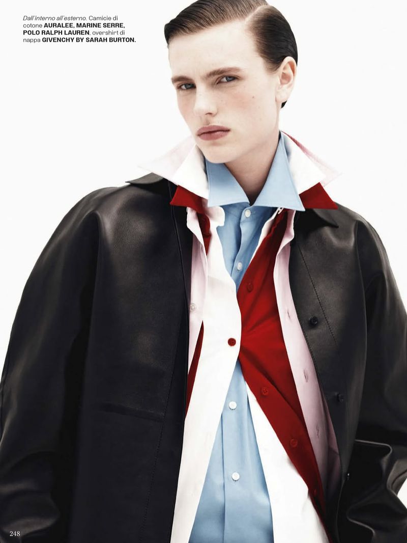 Fashion editorial shot of a young adult model wearing a black leather coat, light blue shirt, and red tie against a plain white backdrop.