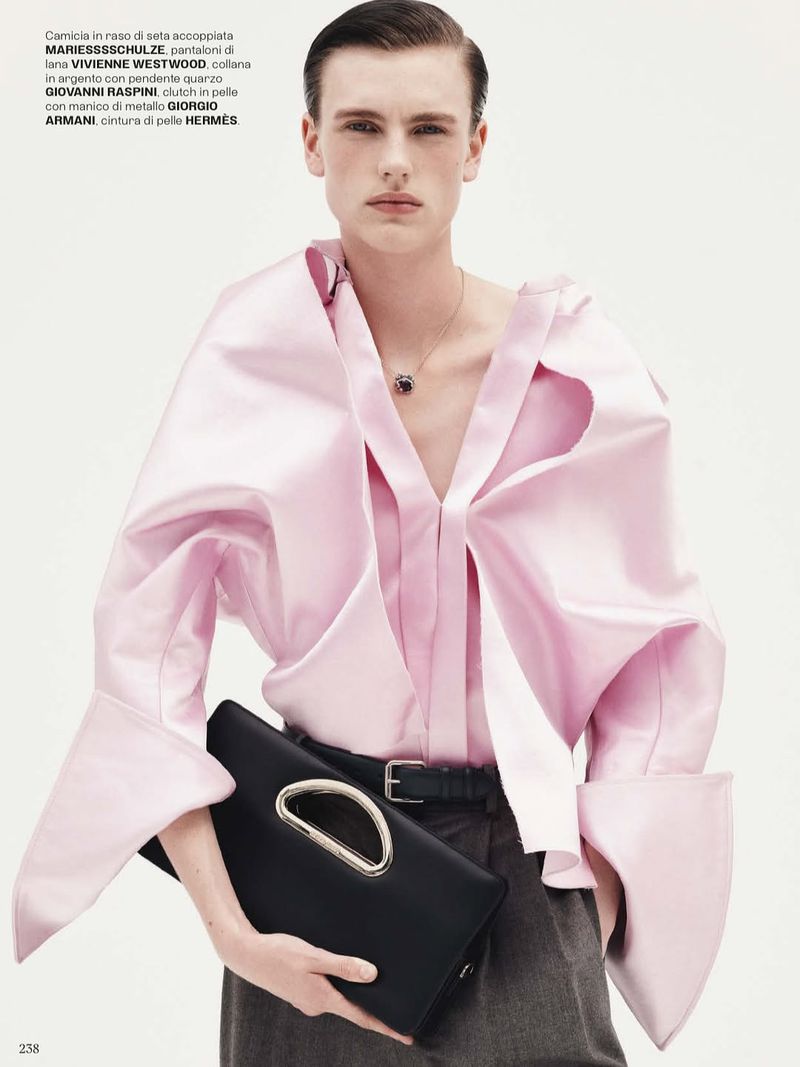 A fair-skinned adult man poses in a light pink, wide-sleeved blouse, holding a black clutch. He wears a necklace, grey pants, and a belt.