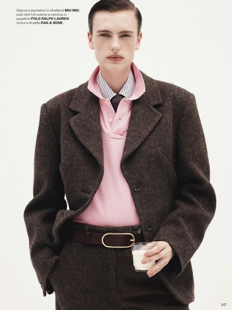 Man poses indoors wearing a brown wool suit, pink polo shirt, striped dress shirt, and tie, holding a glass of milk in a studio fashion shot.