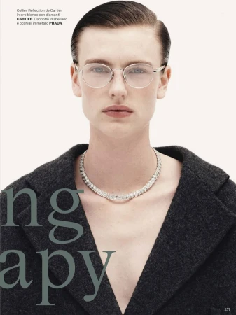 Headshot of a model wearing a gray coat and a diamond necklace looking directly at the camera with round glasses on a neutral background.