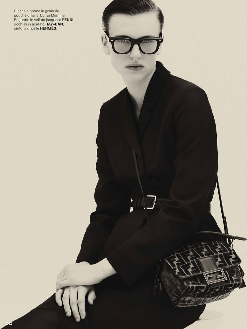 Model in dark glasses and black suit jacket holding a jacquard handbag against a plain background