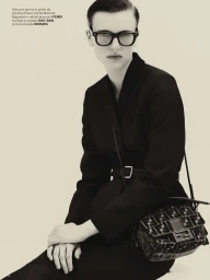 Model in dark glasses and black suit jacket holding a jacquard handbag against a plain background