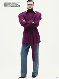 Model wearing a structured purple belted coat and blue jeans standing against a plain white background.