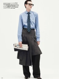 Model in blue button-down shirt and grey pleated skirt holding a newspaper clutch against white background
