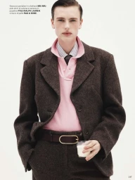 Model wearing a brown tweed jacket and trousers with a pink polo shirt and glass of milk against a white background