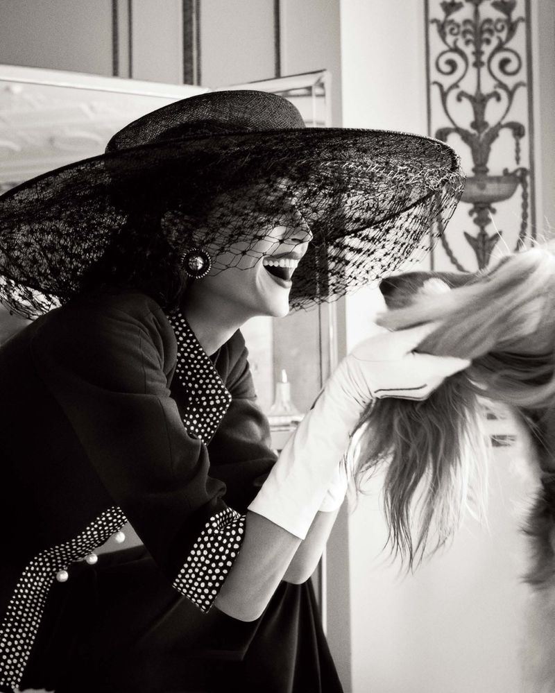 Laughing woman in large veiled hat interacts with a dog, vintage black-and-white style with elegant indoor setting, light background.