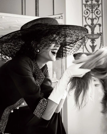Laughing woman in large veiled hat interacts with a dog, vintage black-and-white style with elegant indoor setting, light background.