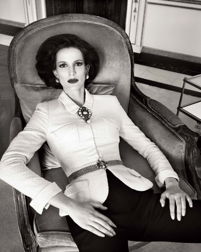 Monochrome portrait of a stylish adult woman with dark hair, wearing a light tweed jacket and dark pants, seated in an armchair.