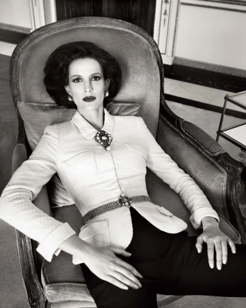 Monochrome portrait of a stylish adult woman with dark hair, wearing a light tweed jacket and dark pants, seated in an armchair.