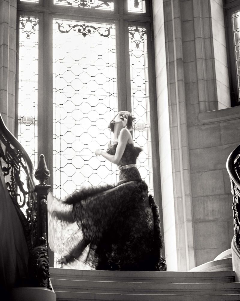 Monochrome portrait of a woman in an elaborate gown descending a grand staircase with large windows, showcasing a blend of fashion and architecture.