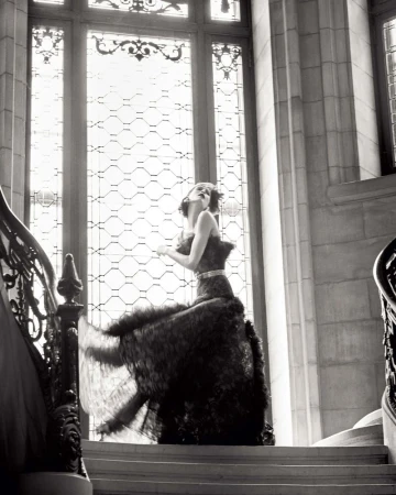 Monochrome portrait of a woman in an elaborate gown descending a grand staircase with large windows, showcasing a blend of fashion and architecture.