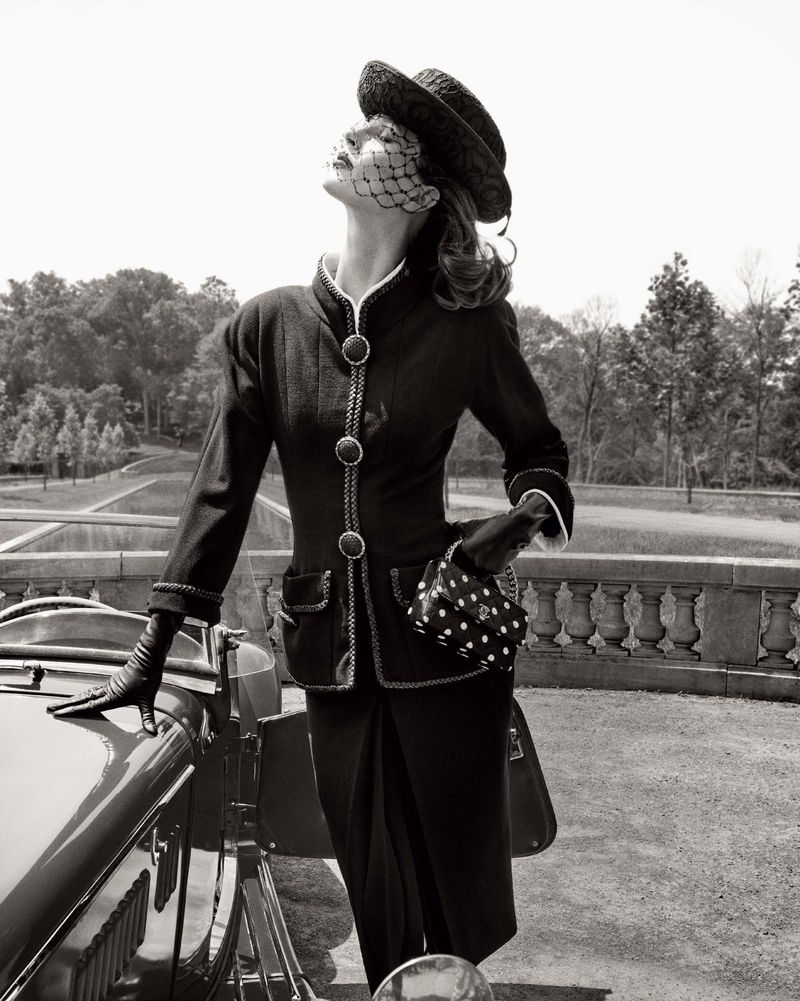 A stylish vintage fashion photograph showcases a woman in a tailored black suit, hat, and gloves, posing next to a classic car in an outdoor setting.
