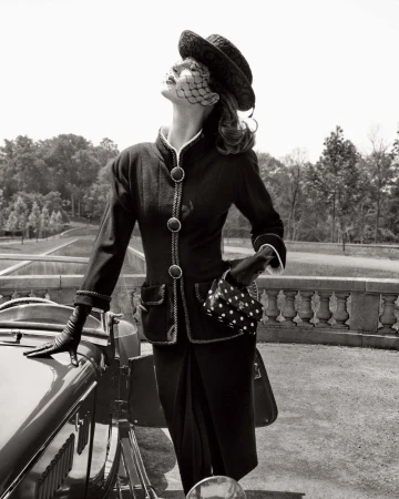 A stylish vintage fashion photograph showcases a woman in a tailored black suit, hat, and gloves, posing next to a classic car in an outdoor setting.