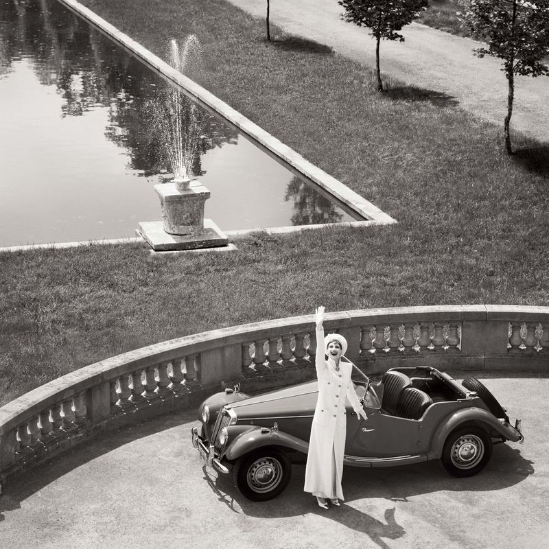 Black and white glamour shot features an elegant woman in a white suit next to a vintage car, with a fountain and manicured lawn.