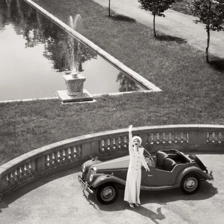 Black and white glamour shot features an elegant woman in a white suit next to a vintage car, with a fountain and manicured lawn.
