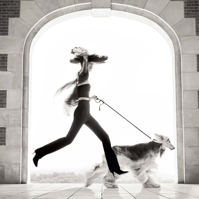 Fashion shot of an adult female in sleek clothing walking an Afghan hound through a stone archway with a light background.