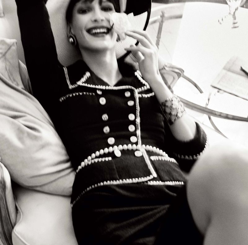 A monochrome portrait of a smiling woman in a pearl-trimmed suit and hat, lying on a cushioned surface, holding a white flower.