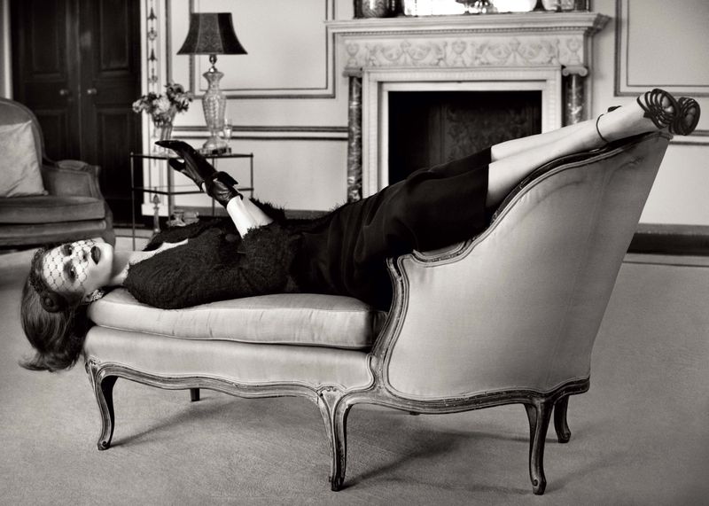 Elegant woman in vintage-style fashion lying on a chaise lounge in a luxurious interior setting with classic design elements.