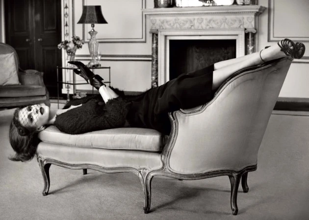 Elegant woman in vintage-style fashion lying on a chaise lounge in a luxurious interior setting with classic design elements.