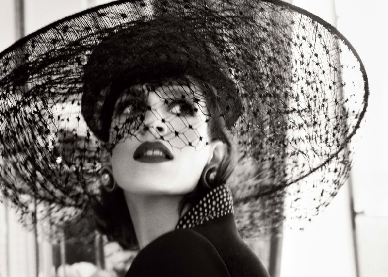 Black-and-white portrait of a woman wearing a large black veiled hat, collar with polka dots, and large earrings, looking upwards.