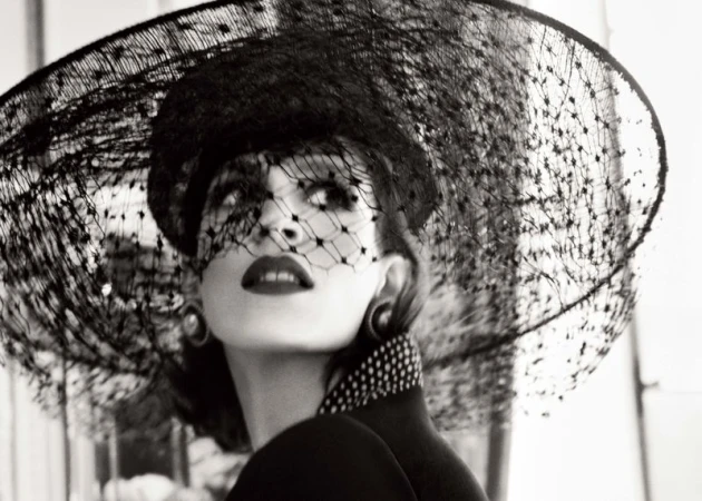 Black-and-white portrait of a woman wearing a large black veiled hat, collar with polka dots, and large earrings, looking upwards.