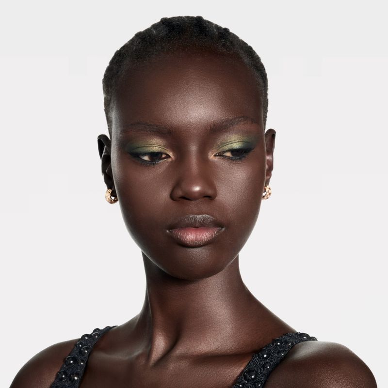 Close-up of a dark-skinned adult female with green eyeshadow and neutral expression, wearing earrings against a white background.