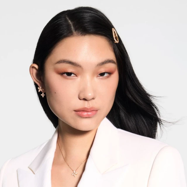 Elegant Asian woman with dark hair in a white blazer poses indoors wearing flower earrings and necklace against a plain background.