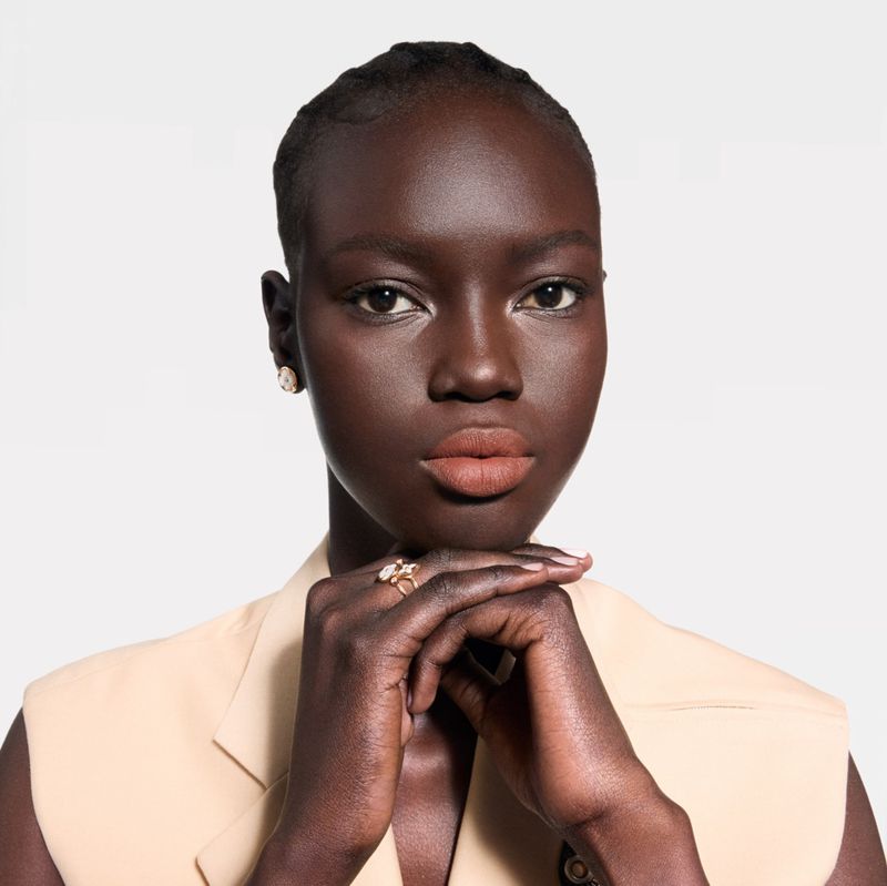 Close-up studio shot of a dark-skinned adult woman with neutral makeup, wearing a cream sleeveless jacket and jewelry against a white backdrop.