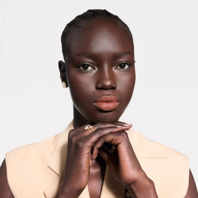 Close-up studio shot of a dark-skinned adult woman with neutral makeup, wearing a cream sleeveless jacket and jewelry against a white backdrop.