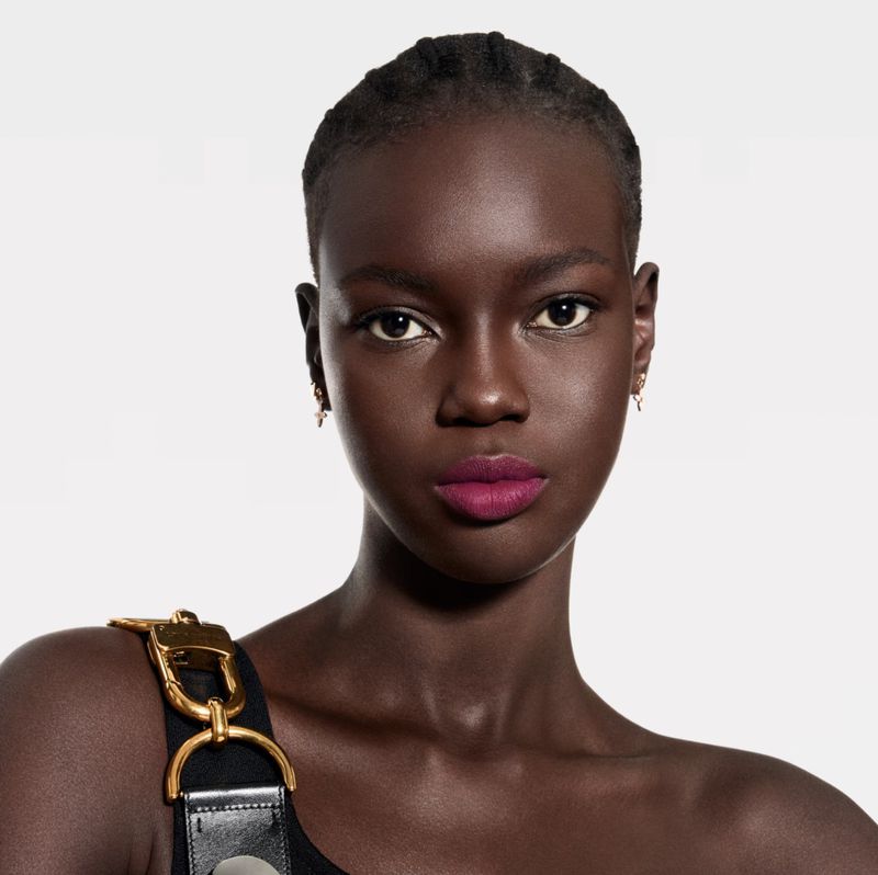 Close-up portrait of an African American woman wearing dark lipstick, gold earrings, and a black tank top with gold hardware against white.