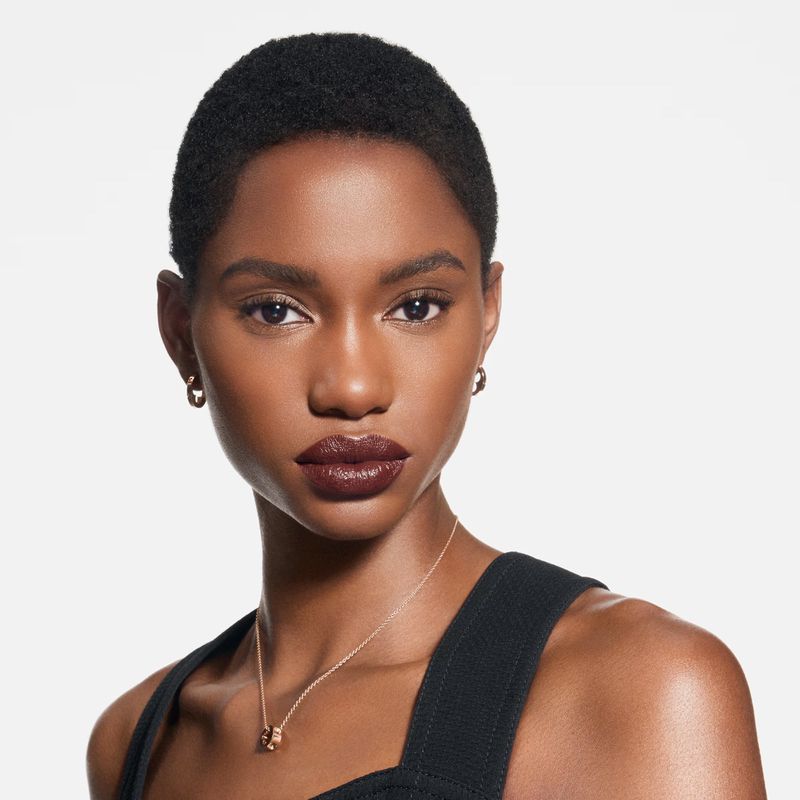 Close-up studio portrait of a dark-skinned woman with short hair wearing dark lipstick, gold jewelry, and a black top against a white backdrop.