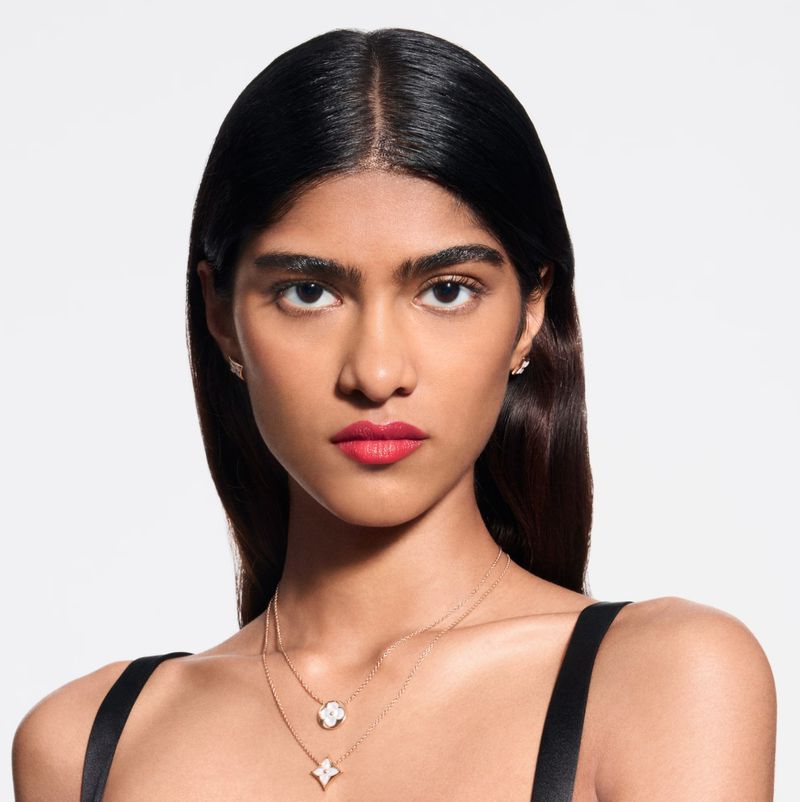Portrait of a woman with fair skin and dark hair. She is wearing lipstick and two layered necklaces. White studio backdrop.