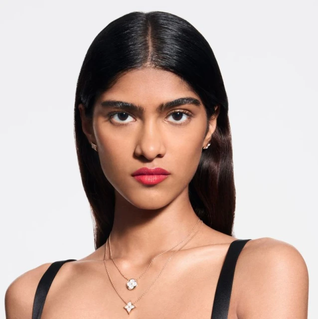 Portrait of a woman with fair skin and dark hair. She is wearing lipstick and two layered necklaces. White studio backdrop.