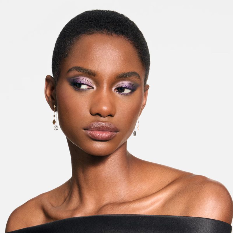 Close-up shot of a Black woman with a short hairstyle, wearing off-the-shoulder black top and sparkling earrings, wearing purple eyeshadow.