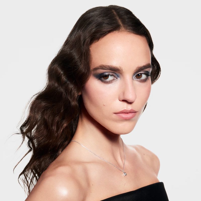 Portrait of a young woman with dark wavy hair and dark blue eye makeup wearing a strapless black top and a silver necklace indoors.