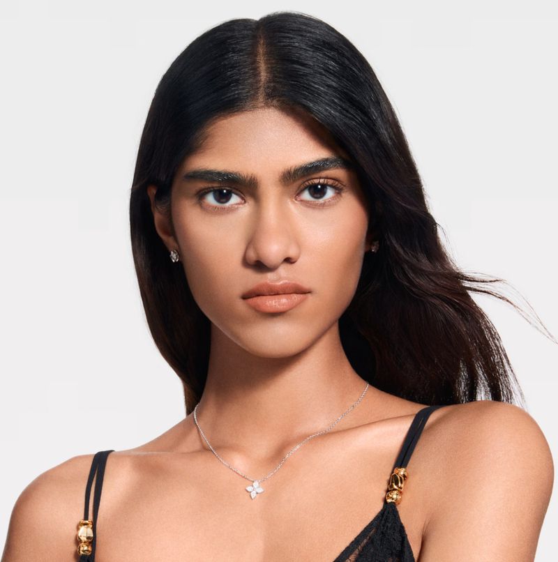 Close-up of a woman with dark hair and neutral makeup wearing a delicate floral pendant necklace and small earrings against a white background.