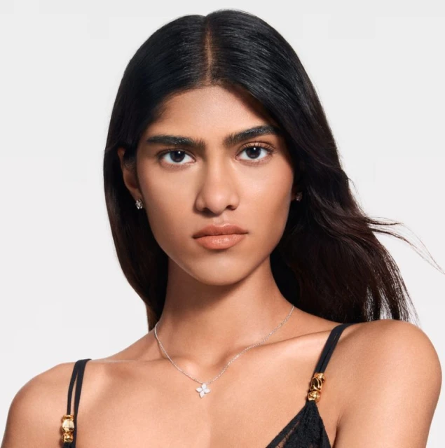 Close-up of a woman with dark hair and neutral makeup wearing a delicate floral pendant necklace and small earrings against a white background.