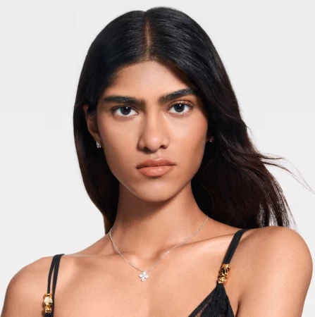 Close-up of a woman with dark hair and neutral makeup wearing a delicate floral pendant necklace and small earrings against a white background.