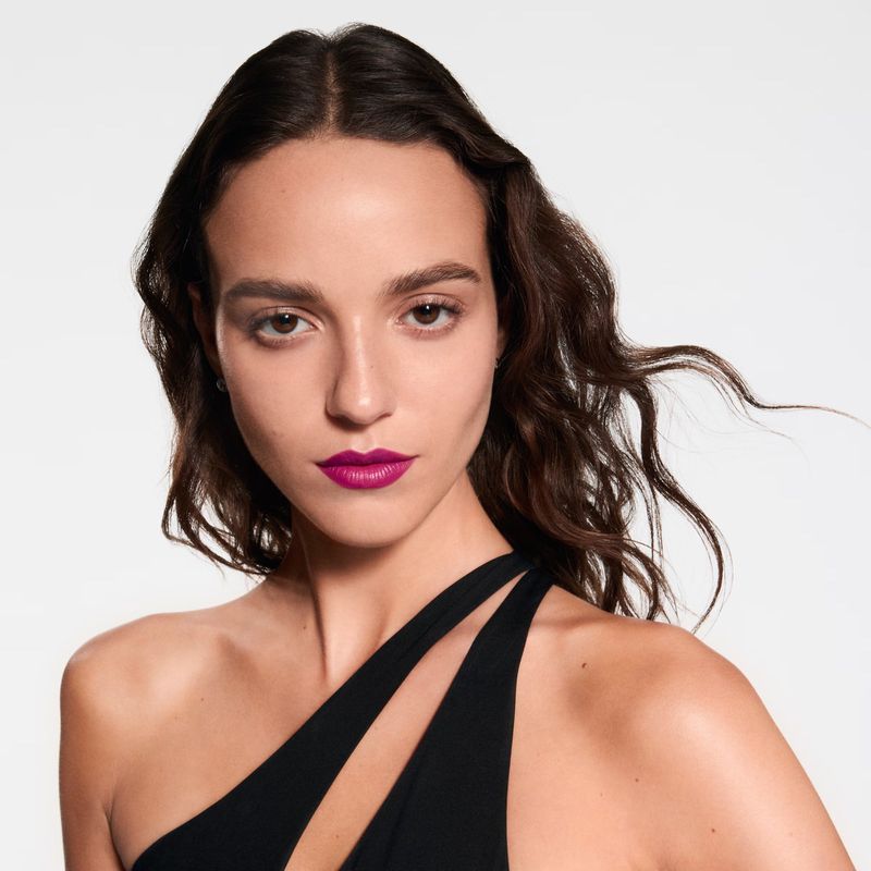 Close-up of a brunette woman with bright pink lipstick, wearing a black, asymmetrical top against a white backdrop in a beauty shot.