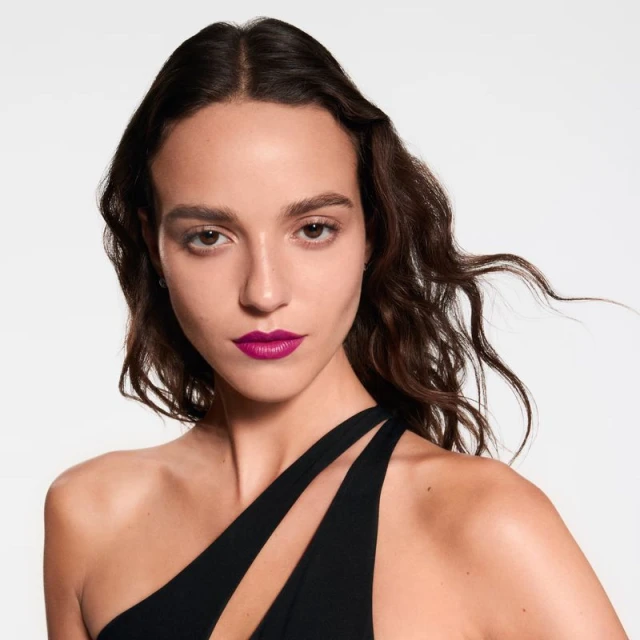 Close-up of a brunette woman with bright pink lipstick, wearing a black, asymmetrical top against a white backdrop in a beauty shot.