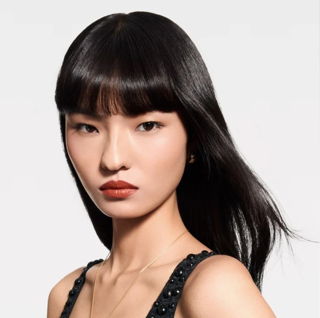 Close-up portrait of a stylish young Asian woman with dark hair, wearing makeup and a beaded dress against a neutral backdrop.