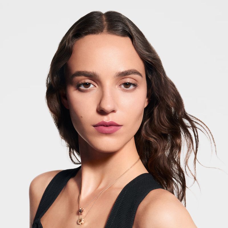 Close-up studio portrait of a young adult woman with long brown hair, wearing a black top and a gold pendant necklace, against white.