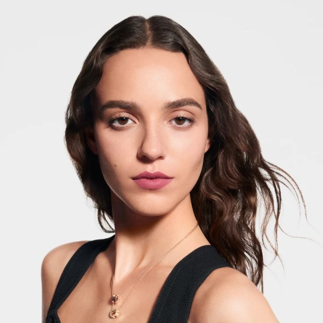 Close-up studio portrait of a young adult woman with long brown hair, wearing a black top and a gold pendant necklace, against white.